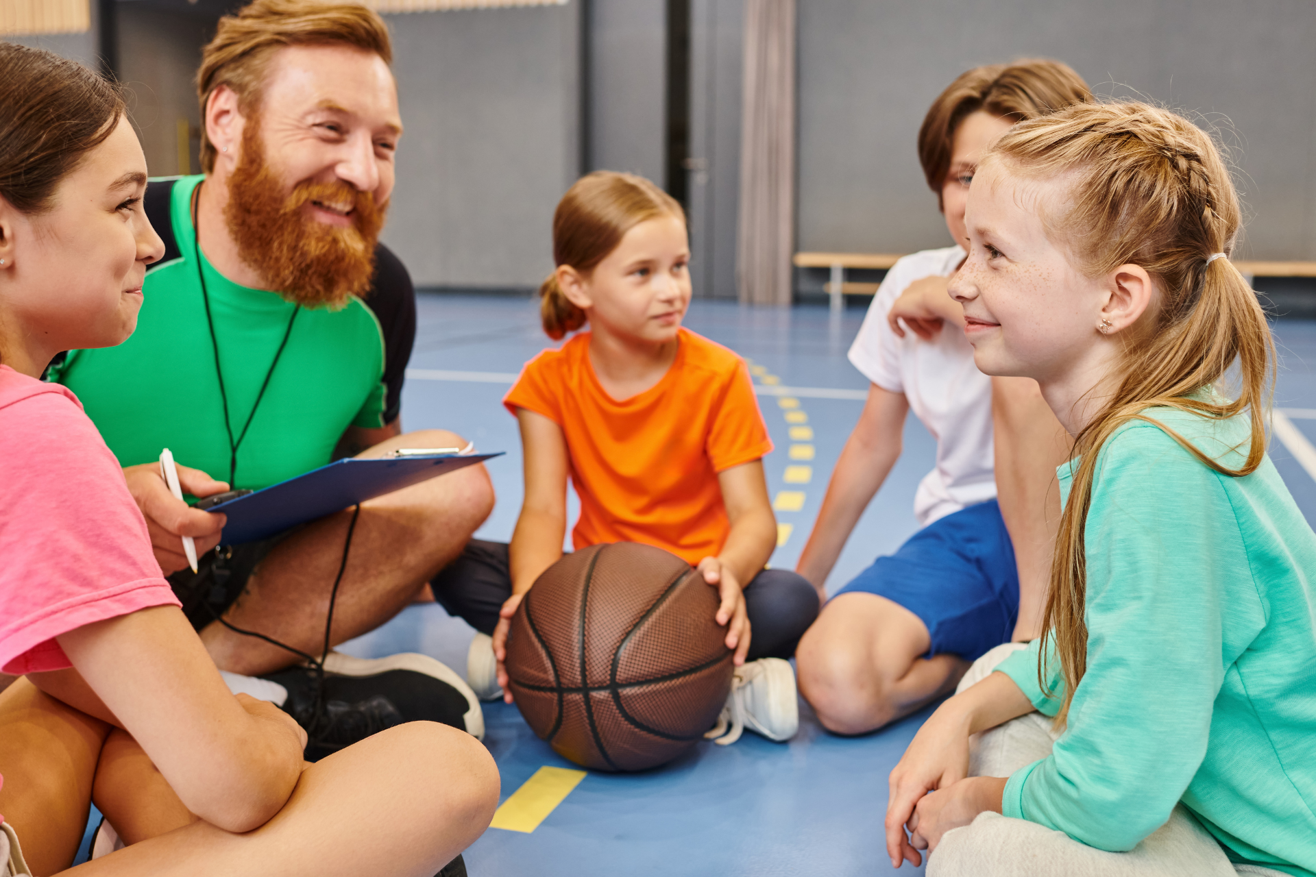students sitting on gym floor with teacher and a basketball